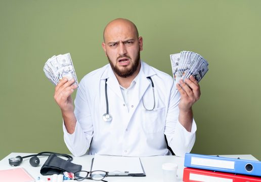 Angry young bald male doctor wearing medical robe and stethoscope sitting at desk