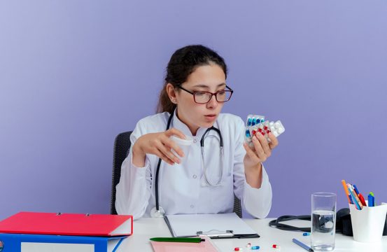 Impressed young female doctor wearing medical robe and stethoscope sitting at desk with medical tools holding and looking at medical pills and holding beaker isolated