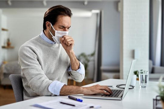 Young entrepreneur coughing while wearing protective face mask and working on a computer at home