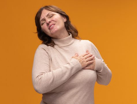 Middle aged woman in beige turtleneck holding hand on her chest feeling thankful with closed eyes standing over orange wall