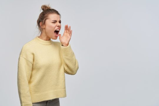 Portrait of angry, adult girl with blond hair gathered in bun. wearing yellow sweater. holds palm next to her mouth and shout to the right at copy space. stand isolated over white wall