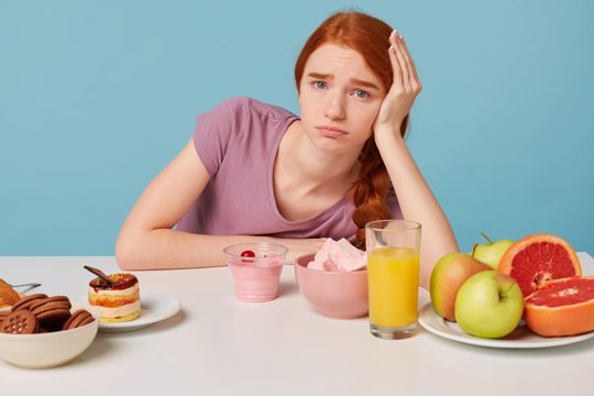 Young red-haired girl sits at the table with her head on her hand sadly understands the importance of fresh fruit