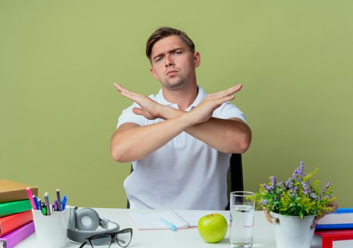 Strict young handsome male student sitting at desk with school tools showing gesture of no on olive green