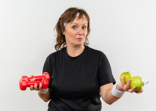 Middle aged sporty woman in black t-shirt with headband holding two green apples and dumbbells looking confused having doubts standing over white wall