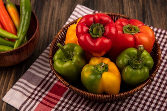 Top view of colorful bell peppers on a bucket on a checked cloth with long shaped green peppers on a bowl on a wooden wall