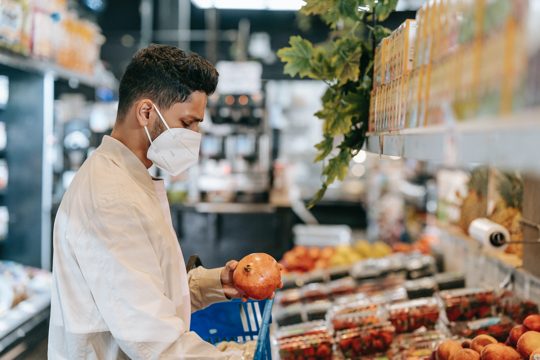 Side view of young Hispanic man in protective mask choosing fresh pomegranate from stall in supermarket during coronavirus pandemic