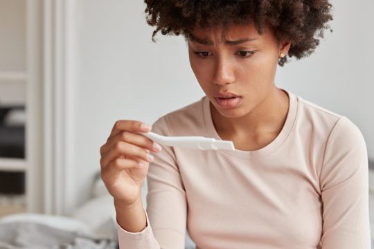 Embrarrassed woman with afro hairstyle faces serious problem, dissatisfied with pregnancy test